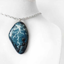 Load image into Gallery viewer, a beach stone with cat art: a painting of a black cat on a cliff, looking out into the night sky, filled with lightning. this piece is available as a keepsake, magnet or a pendant necklace