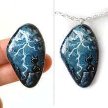 Load image into Gallery viewer, a beach stone with cat art: a painting of a black cat on a cliff, looking out into the night sky, filled with lightning. this piece is available as a keepsake, magnet or a pendant necklace