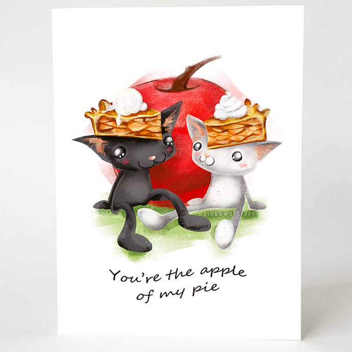 a greeting card with art of a black cat and white cat, both wearing slices of apple pie on their heads. they sit in front of a giant apple. card reads, you're the apple of my pie