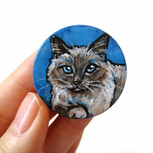 Load image into Gallery viewer, A circle wooden disc, with cat art of a Birman cat with blue eyes, with a blue background. Available as either a wood keepsake, magnet or pendant necklace
