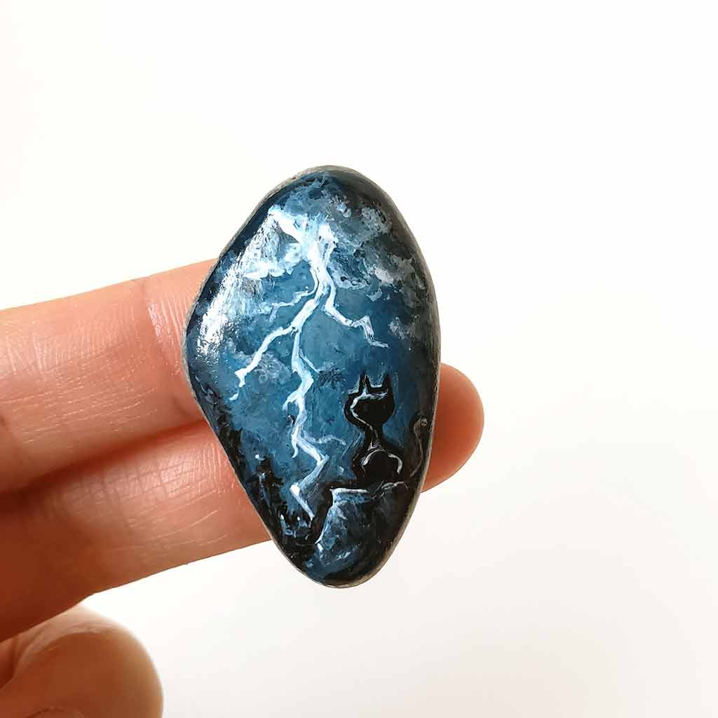 a beach stone with cat art: a painting of a black cat on a cliff, looking out into the night sky, filled with lightning. this piece is available as a keepsake, magnet or a pendant necklace