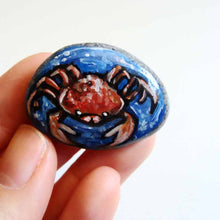 Load image into Gallery viewer, a small beach rock, hand painted with zodiac art: an orange and red crab, the sign of cancer, surrounded by blue water. available as a stone keepsake, magnet or pendant necklace
