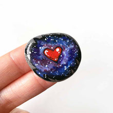 Load image into Gallery viewer, a beach stone has been hand painted with a heart surrounded by outer space and stars, and hand crafted as either a keepsake stone, magnet, or pendant necklace
