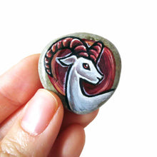 Load image into Gallery viewer, a beach rock painted with the zodiac art of aries: a white ram with dark red horns. this stone is available as a keepsake, magnet or pendant necklace
