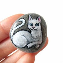 Load image into Gallery viewer, A small beach stone hand painted with a portrait of a white cat with blue eyes, available as a stone, magnet or pendant necklace.
