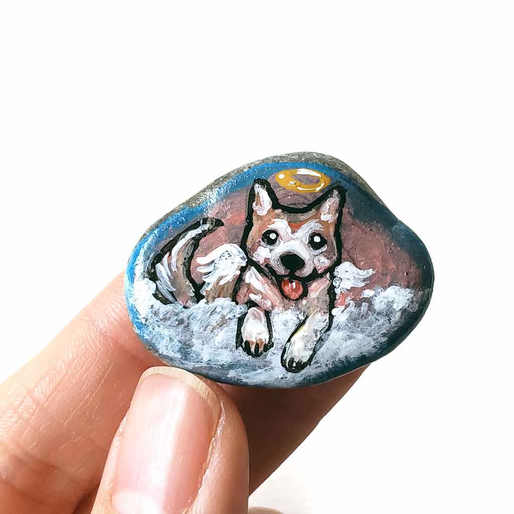 a small smooth beach stone, hand painted with the portrait of an akita dog, painted as an angel, it peers out over fluffy clouds against a blue and pink sky. this piece is available as a keepsake or pendant necklace