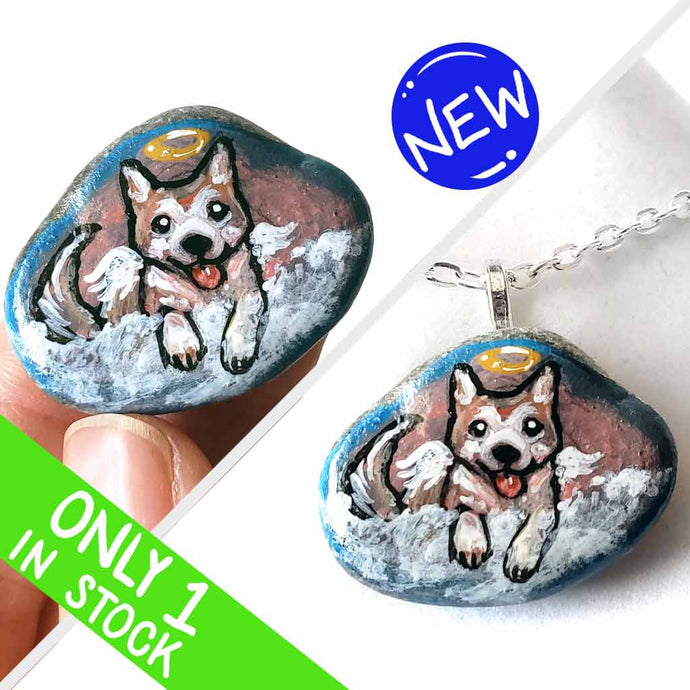 a small smooth beach stone, hand painted with the portrait of an akita dog, painted as an angel, it peers out over fluffy clouds against a blue and pink sky. this piece is available as a keepsake or pendant necklace
