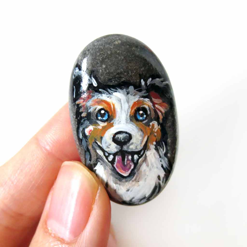 a small beach rock, hand painted with art of a smiling Australian Shepherd dog, available as a stone keepsake, fridge magnet, or pendant necklace. 
