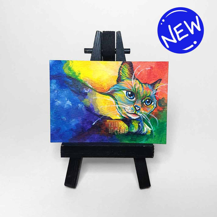 an aceo sized painting featuring a birman cat painted with rainbow colours. the painting sits on a tiny black easel