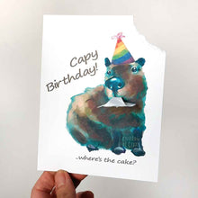 Load image into Gallery viewer, a greeting card featuring an illustration of a capybara wearing a rainbow birthday hat. The top right corner of the card hangs from its mouth. the front reads, Capy Birthday! ..where&#39;s the cake?
