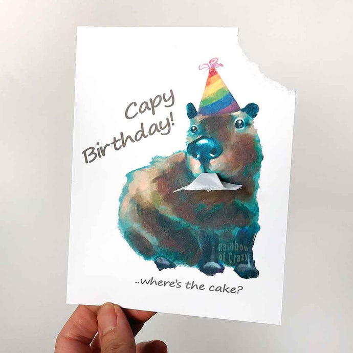 a greeting card featuring an illustration of a capybara wearing a rainbow birthday hat. The top right corner of the card hangs from its mouth. the front reads, Capy Birthday! ..where's the cake?