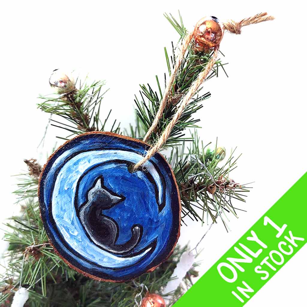 a wood slice ornament, hand painted with the silhouette of a black cat, curled up on a crescent moon, against a dark blue background