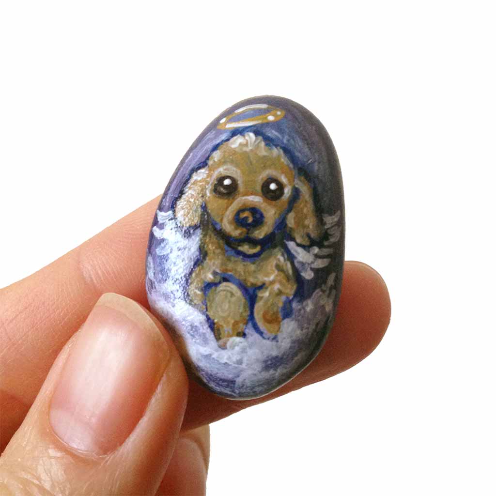 a small beach stone hand painted with the portrait of a brown poodle as an angel, sitting in the clouds against a blue sky. available as a keepsake or pendant necklace