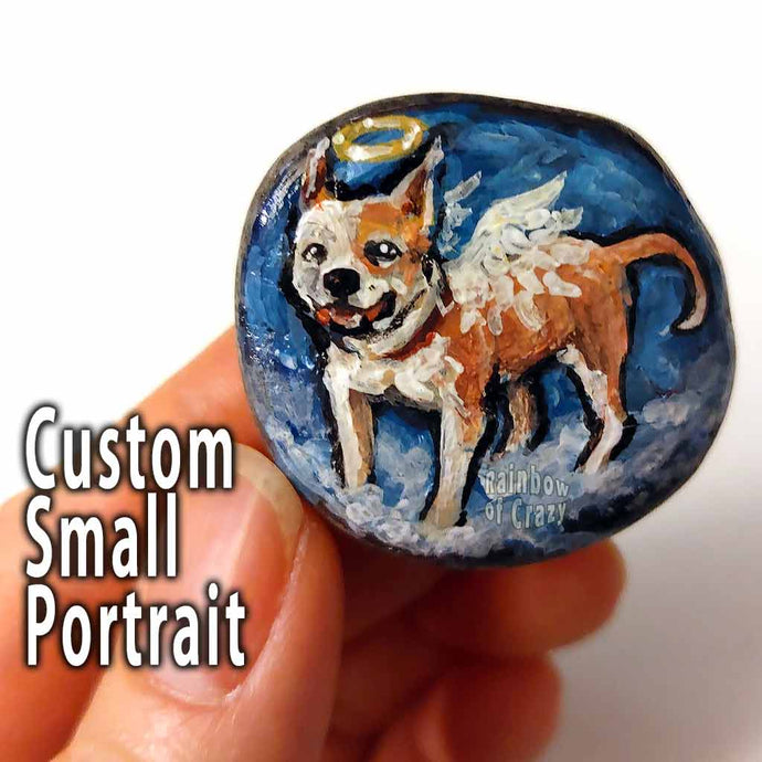 hand holding a small beach stone, hand painted with a portrait of a brown and white dog, painted as an angel with halo and wings, standing on clouds against a blue sky.