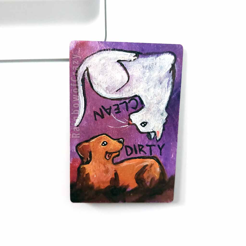 wood magnet on a dishwasher, hand painted with a white cat with the word CLEAN next to it. at the top, upside down, is a Golden labrador dog that is covered in mud, with word DIRTY next to it, background a purple colour