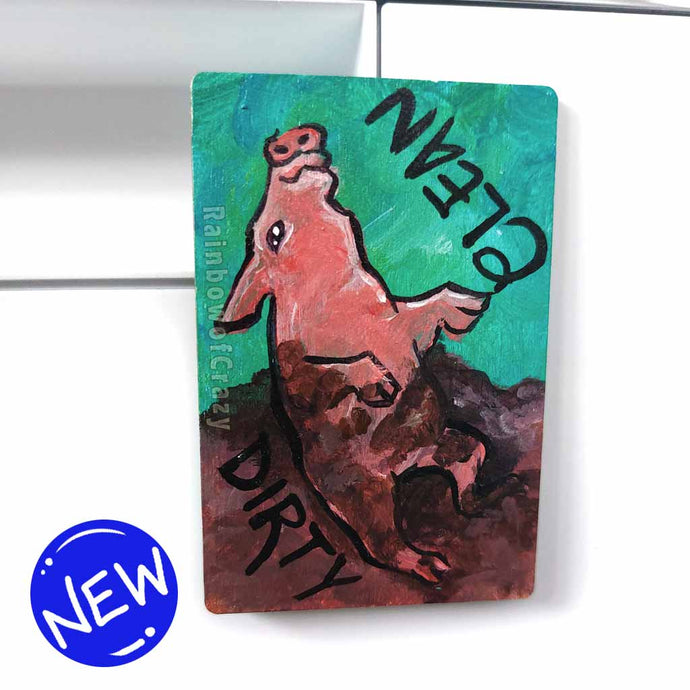 wood magnet on a dishwasher, hand painted with a pig with the word CLEAN (upside down) next to its face at the top. the second half of the pig is muddy, and written next to its body, DIRTY.