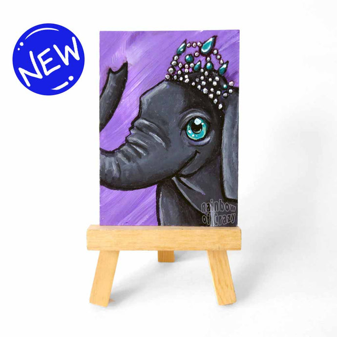 an aceo sized painting on a wood easel, featuring an elephant wearing a tiara, painted against a soft purple background