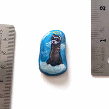 Load image into Gallery viewer, a small beach stone, painted with a smiling ferret angel, with halo and wings, on top of clouds, against a blue sky. available as a keepsake, magnet or pendant necklace
