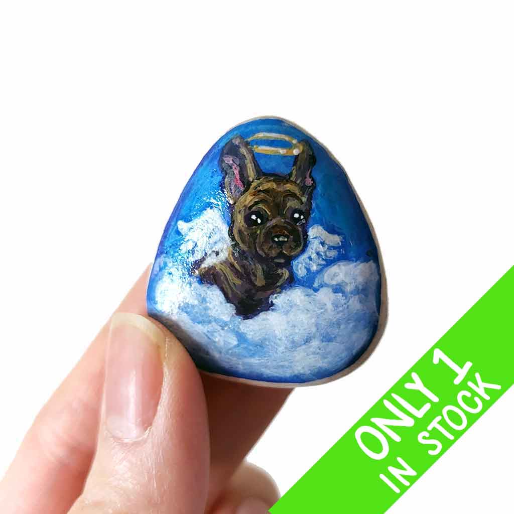 a small beach stone, hand painted with a portrait of a brindle french bulldog, as an angel in the clouds.