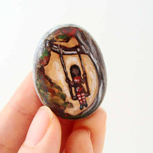 Load image into Gallery viewer, small rock art: a beach stone is hand painted with a girl in red, sitting on a tree swing, available as either a keepsake, fridge magnet, or a necklace
