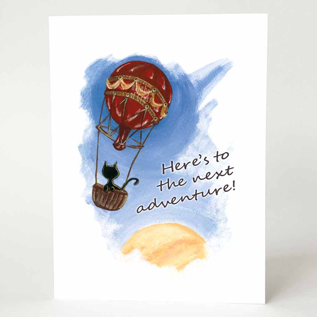 a greeting card with an illustration of a black cat in a hot air balloon, floating in the sky with the sun partially in view on the bottom right. the front of the card reads 