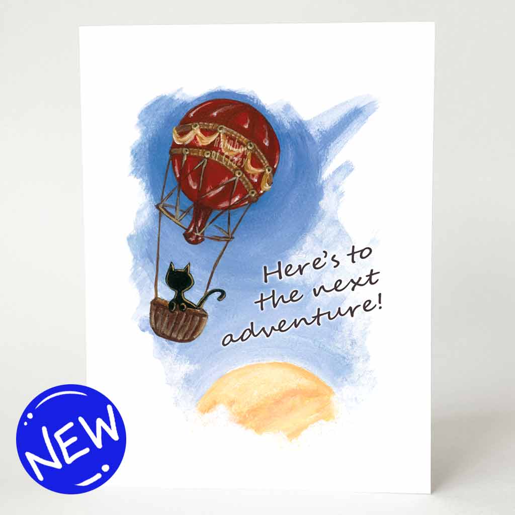 a greeting card with an illustration of a black cat in a hot air balloon, floating in the sky with the sun partially in view on the bottom right. the front of the card reads "Here's to the next adventure!"