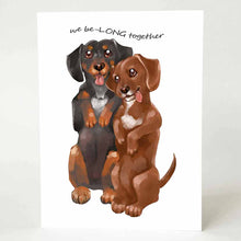 Load image into Gallery viewer, a greeting card with an illustration of one black dachshund, and one brown dachshund, both standing up on their hind legs. it reads, &quot;we be-LONG together&quot;
