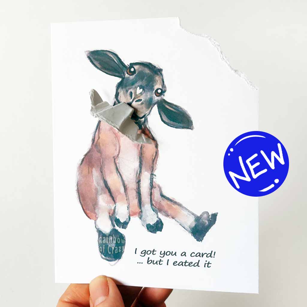 a funny greeting card with a black and brown sheep sitting upright, with the top right corner of the card ripped and hanging from its mouth. the card reads, 