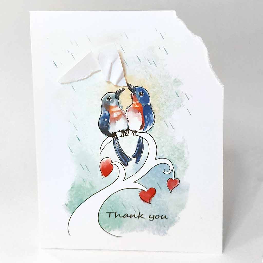 a handmade greeting card wtih an illustration of two bluebirds on a tree branch. the top right corner of the card is ripped off and hangs from one bluebird's mouth, as it holds it up above the other bird, protecting it from the rain. the card says, 