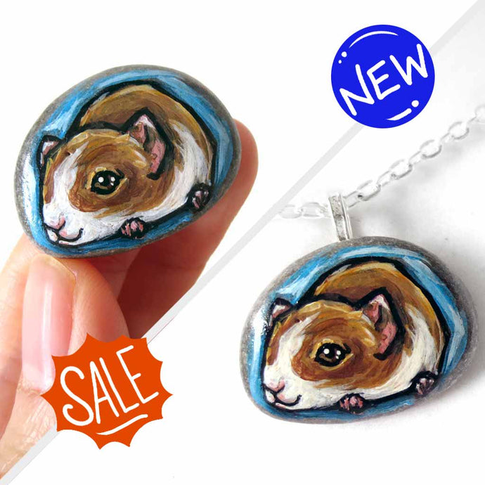 a small beach stone hand painted with a portrait of a guinea pig against a blue background. Available as a keepsake stone or pendant necklace