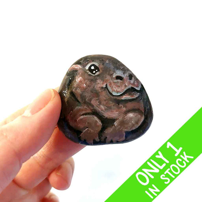 a hand holding a smooth, flat, beach stone hand painted with a smiling hippo