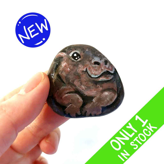 a hand holding a smooth, flat, beach stone hand painted with a smiling hippo