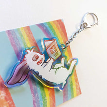 Load image into Gallery viewer, A clear, rainbow holographic acrylic keychain featuring a character from Rainbow of Crazy Comics: a smiling mini lop rabbit lounges on his back as he reads a book with a picture of a rabbit on the back.
