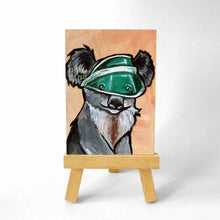 Load image into Gallery viewer, an aceo sized painting of a smiling koala wearing a green sun visor, against an orange background. the painting sits on a small wood easel
