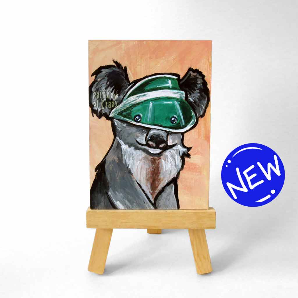 an aceo sized painting of a smiling koala wearing a green sun visor, against an orange background. the painting sits on a small wood easel