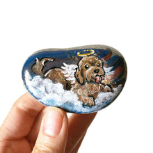 Load image into Gallery viewer, a smooth beach stone featuring rock art of a labradoodle dog, painted as an angel in the clouds
