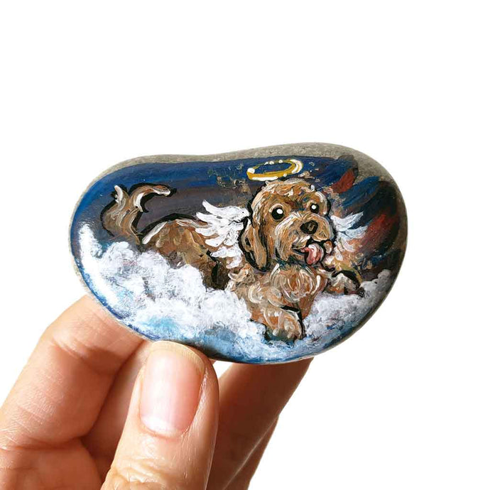 a smooth beach stone featuring rock art of a labradoodle dog, painted as an angel in the clouds