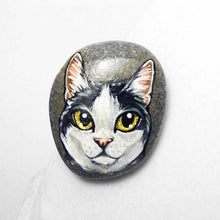 Load image into Gallery viewer, a smooth beach stone, painted with a close-up portrait of a black and white manx cat with yellow eyes and pink ears.
