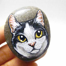 Load image into Gallery viewer, a smooth beach stone, painted with a close-up portrait of a black and white manx cat with yellow eyes and pink ears.
