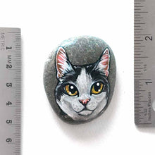 Load image into Gallery viewer, a smooth beach stone, painted with a close-up portrait of a black and white manx cat with yellow eyes and pink ears.
