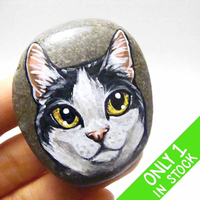 a smooth beach stone, painted with a close-up portrait of a black and white manx cat with yellow eyes and pink ears.