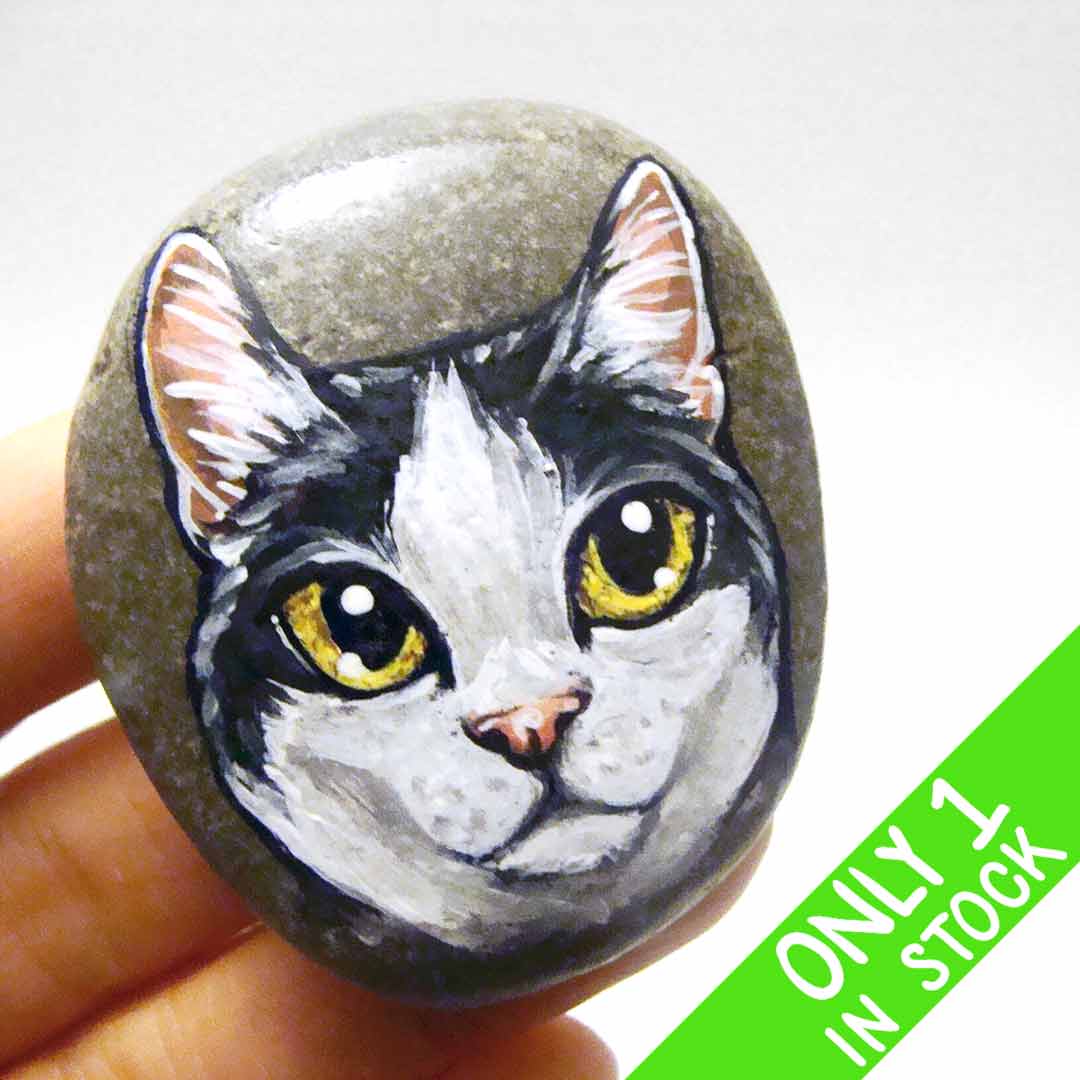 a smooth beach stone, painted with a close-up portrait of a black and white manx cat with yellow eyes and pink ears.