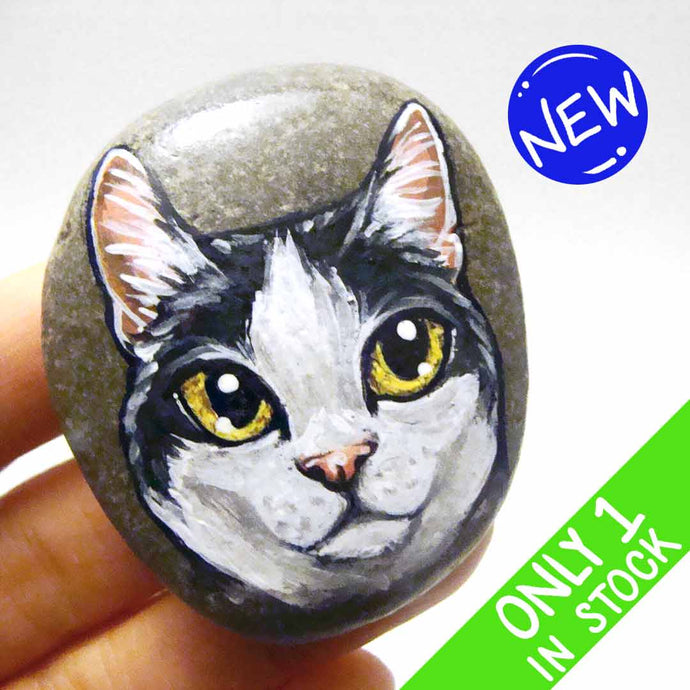 a smooth beach stone, painted with a close-up portrait of a black and white manx cat with yellow eyes and pink ears.