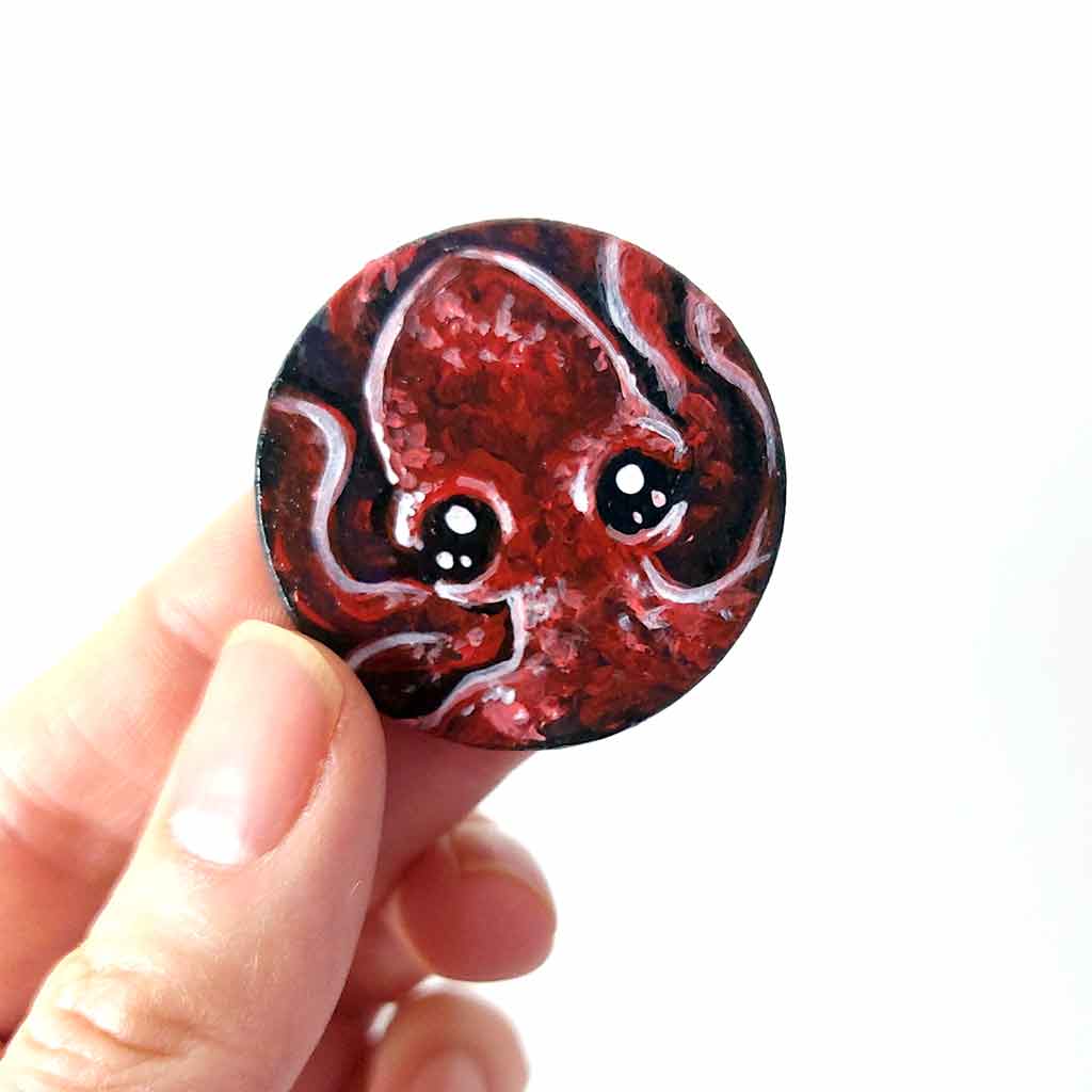 a lightweight wood disc, hand painted with a portrait of a red baby octopus. the painting is available as a keepsake, or pendant necklace