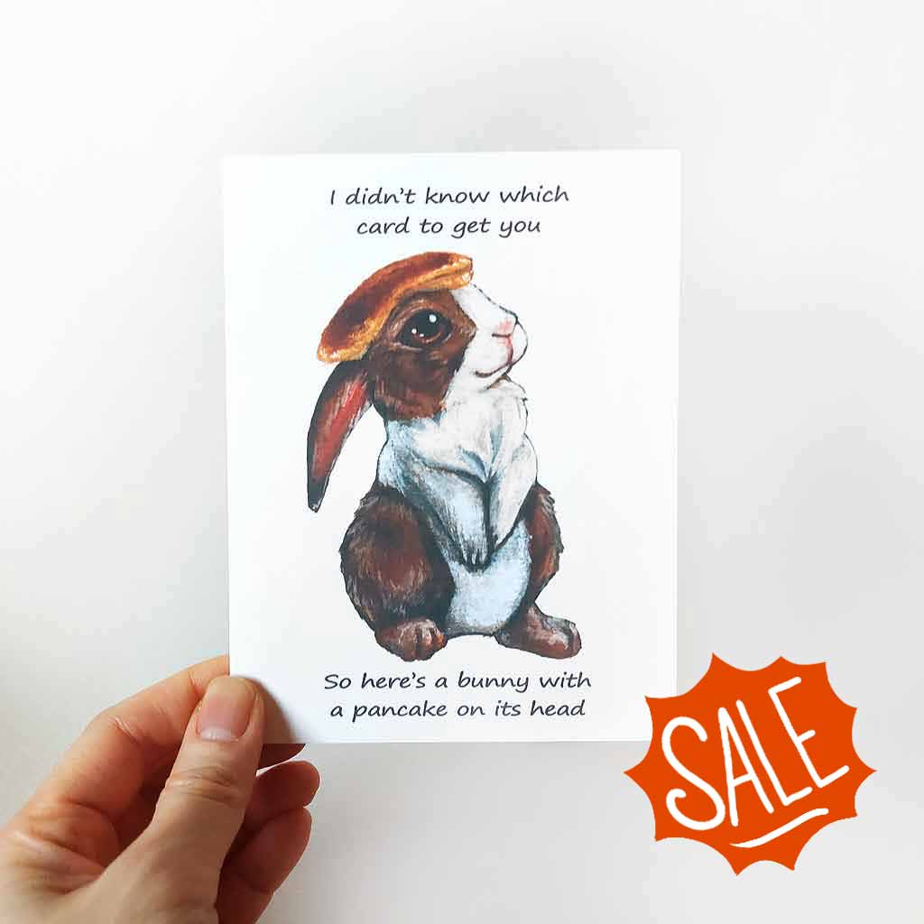 a greeting card, featuring an illustration of a brown and white rabbit with a pancake on its head. front reads, 