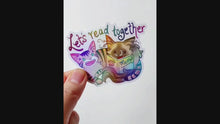 Load and play video in Gallery viewer, a hand holding a sticker with a tabby cat next to a ragdoll cat that's holding a book titled "Meow". At the top of the sticker, it says, "Let's read together". the hand tilts the sticker slightly to show the holographic finish
