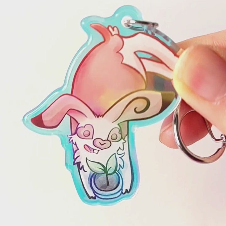 A clear, blue-tinted holographic acrylic keychain featuring a character from Rainbow of Crazy Comics: an English spot rabbit, lying down, admires a potted plant. 