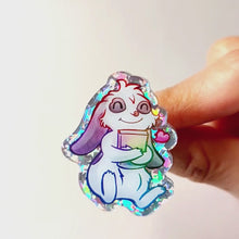 Load and play video in Gallery viewer, acrylic glitter pin featuring an illustration of a mini lop rabbit hugging a book, with a tiny heart next to him.
