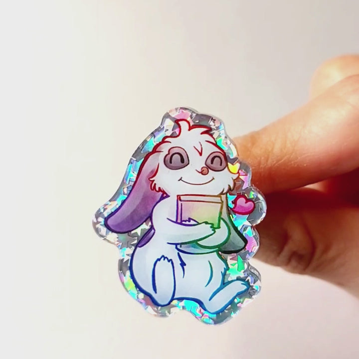 acrylic glitter pin featuring an illustration of a mini lop rabbit hugging a book, with a tiny heart next to him.