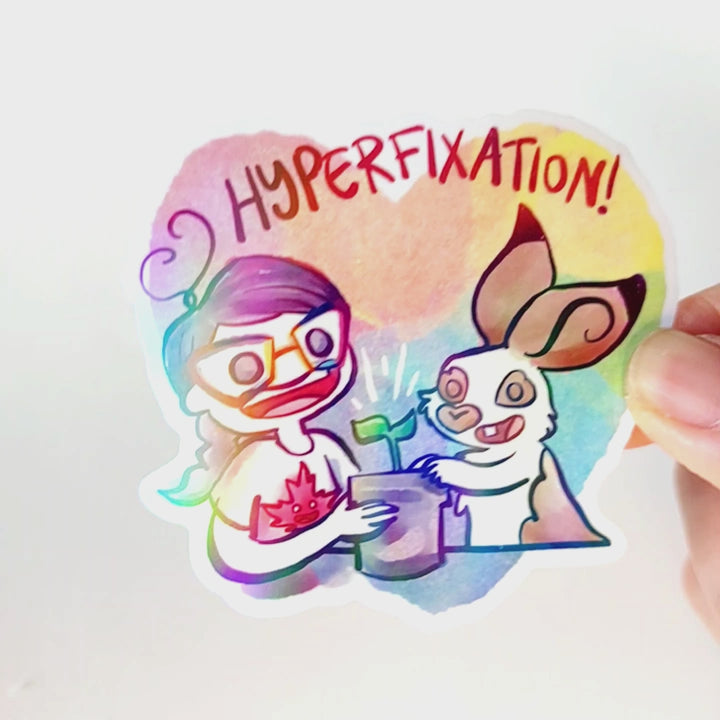Heart shaped holographic sticker with the word "HYPERFIXATION!" above a Comic Jo and an English Spot rabbit, both smiling as they tend to a potted plant.